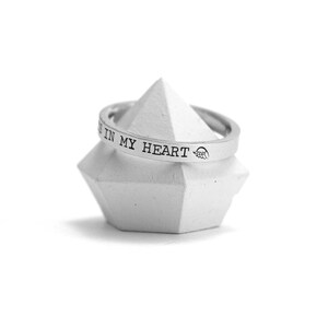 Op de afbeelding: Een zilveren ring met de inscriptie "You are in my heart" op een witte displaystandaard.