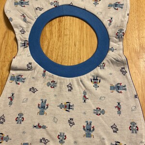 Op de afbeelding: Een baby slabbetje met een lichtgrijze achtergrond en een patroon van kleurrijke robots. De slab heeft een blauwe halsband en een afgeronde vorm. De robots zijn in verschillende poses en kleuren, waaronder blauw, rood en geel.