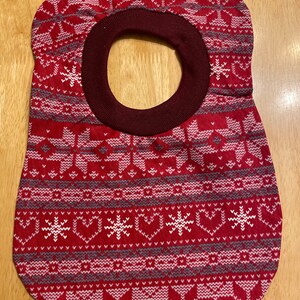 May include: Red and grey patterned fabric bib with a maroon neck opening. The bib has a snowflake and heart design.