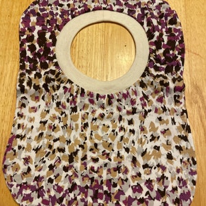 May include: A white bib with a brown, purple, and gray abstract print. The bib has a white, ribbed neck opening.