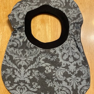May include: A grey fabric bib with a black trim and a damask pattern. The bib has a large opening in the centre.