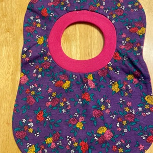 May include: Purple fabric baby bib with a pink trim and a floral pattern of small white, yellow, red, and pink flowers.