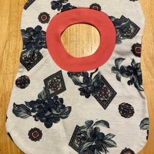 May include: A white baby bib with a floral pattern in shades of blue and grey. The bib has a pink trim around the neck opening.