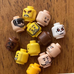 May include: A collection of 12 yellow, brown, and white Lego minifigure heads with various facial expressions. The heads are arranged in a circle on a wooden surface. A white note with the number "#1" written in blue ink is in the lower right corner of the image.