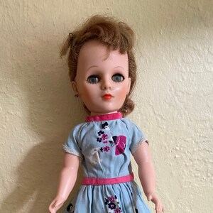 May include: A vintage plastic doll with brown hair, blue eyes, and a light blue dress with pink and white floral accents.