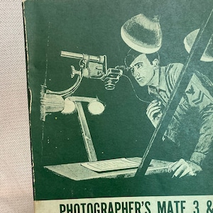 May include: A green cover of a training manual for the United States Navy. The cover features a black and white illustration of a sailor looking through a camera lens. The title of the manual is "Photographer's Mate 3 & 2". The text "Bureau of Naval Personnel" and "Rate Training Manual" are also visible. The text "NAVPERS 10355-A" is printed in the bottom right corner.