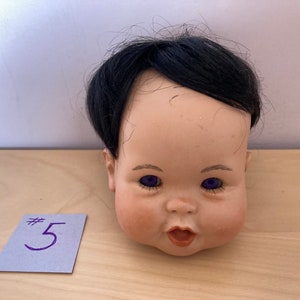 May include: A porcelain doll head with black hair and purple eyes. The doll's mouth is open and it has a rosy complexion. The doll head is sitting on a wooden surface with a blue note card that says #5.