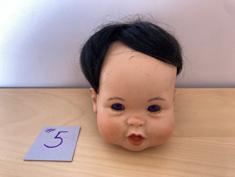 May include: A porcelain doll head with black hair and purple eyes. The doll's mouth is open and it has a rosy complexion. The doll head is sitting on a wooden surface with a blue note card that says #5.
