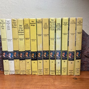 May include: A collection of 11 Nancy Drew mystery books with yellow covers and black text. The books are arranged in a row on a wooden surface. The titles of the books include "The Mystery at Lilac Inn", "The Secret of Red Gate Farm", "The Clue in the Diary", "The Clue of the Broken Locket", "The Whispering Statue", "The Haunted Bridge", "The Clue of the Tapping Heels", "The Mystery of the Brass Bound Trunk", "The Secret in the Old Attic", "The Clue in the Crumbling Wall", "The Invisible Intruder", and "The Double Jinx Mystery".