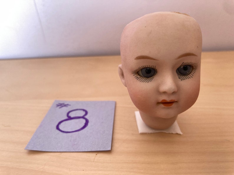 May include: A porcelain doll head with painted eyes and a closed mouth. The doll head is white with a light pink blush on the cheeks. The doll has dark brown painted eyebrows and eyelashes.
