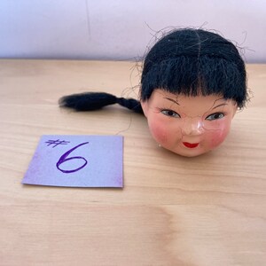 May include: A doll head with black hair and a painted face. The doll head is on a wooden surface with a purple note that says #6.