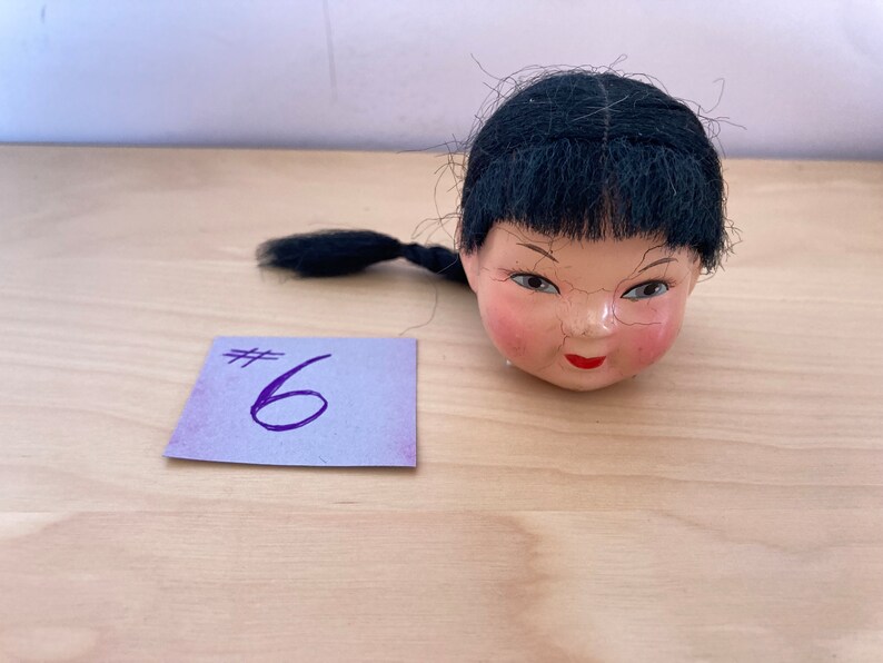 May include: A doll head with black hair and a painted face. The doll head is on a wooden surface with a purple note that says #6.