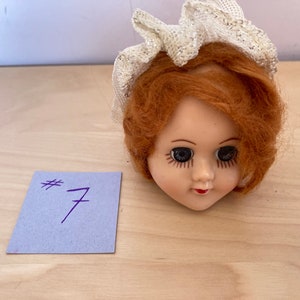 May include: A doll head with red hair and a white lace hat. The doll has blue eyes and a painted smile. The doll head is on a light wood surface.