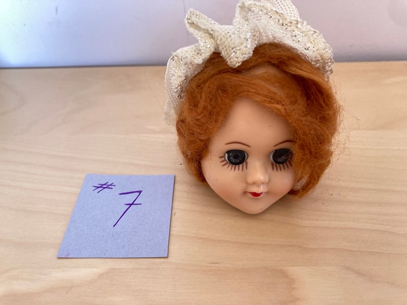 May include: A doll head with red hair and a white lace hat. The doll has blue eyes and a painted smile. The doll head is on a light wood surface.