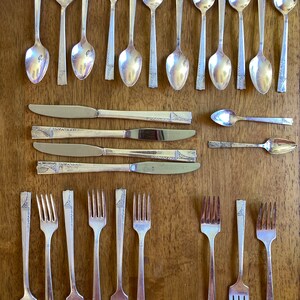 May include: An assortment of vintage silverware, including spoons, knives, and forks, arranged on a wooden surface. The silverware has a silver finish with decorative detailing on the handles. The spoons have a rounded bowl shape, and the knives have a blade. The forks have four tines.