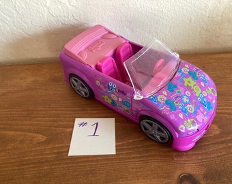 May include: A purple toy convertible car with pink interior and silver wheels. The car has a clear windshield and is decorated with colorful floral and abstract designs. The word "Polly" is visible on the car.