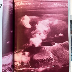 Puede incluir: Una vista a&eacute;rea de un cr&aacute;ter volc&aacute;nico con una columna de humo que se eleva desde el centro. El cr&aacute;ter est&aacute; rodeado por un anillo de monta&ntilde;as y el paisaje es de un color rojo intenso.