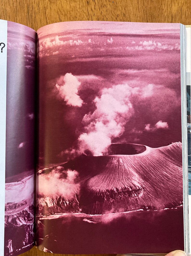 Puede incluir: Una vista a&eacute;rea de un cr&aacute;ter volc&aacute;nico con una columna de humo que se eleva desde el centro. El cr&aacute;ter est&aacute; rodeado por un anillo de monta&ntilde;as y el paisaje es de un color rojo intenso.