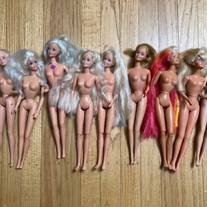 May include: A collection of nine Barbie dolls with long blonde hair, some with pink and yellow highlights. The dolls are all nude and have blue eyes. They are arranged in a row on a wooden surface.