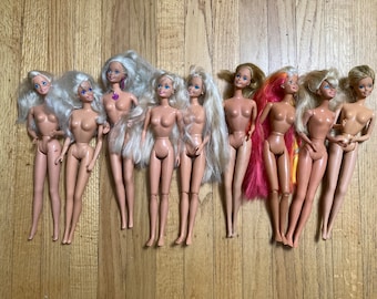May include: A collection of nine Barbie dolls with long blonde hair, some with pink and yellow highlights. The dolls are all nude and have blue eyes. They are arranged in a row on a wooden surface.