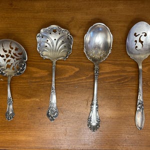 May include: Four antique silver serving spoons with ornate handles and decorative bowls. The spoons include a slotted spoon, a spoon with a floral design, and two solid spoons. The spoons are arranged on a wooden surface.
