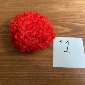 May include: A bright red curly wig for dolls. The wig is on a wooden surface with a white note that says #1.
