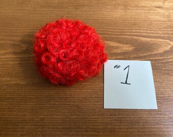 May include: A bright red curly wig for dolls. The wig is on a wooden surface with a white note that says #1.