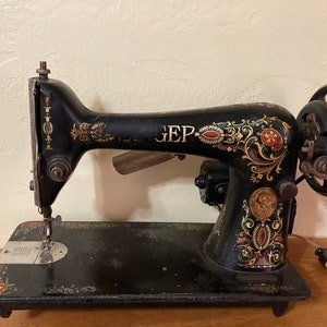 May include: Antique black sewing machine with gold and red floral accents. The machine features the letters "EP" and a hand wheel. The sewing machine is on a wooden surface.