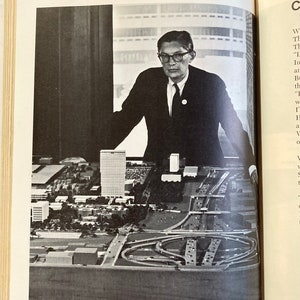 Puede incluir: Una foto en blanco y negro de un arquitecto mirando una maqueta de una ciudad. La maqueta incluye edificios, carreteras y una gran carretera circular.