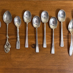 May include: A collection of antique silverware, including spoons and a fork, arranged on a wooden surface. The spoons vary in design, with ornate and simple handles. The fork and spoons are made of a shiny, silver-colored metal.