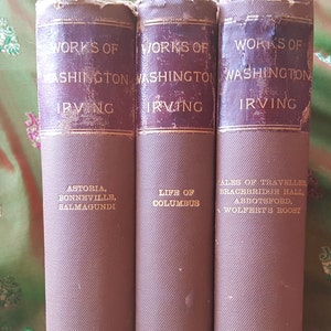 May include: Three brown hardcover books with gold lettering. The books are titled "Works of Washington Irving" and each book lists different works by the author. The first book lists "Astoria, Bonneville, Salmagundi". The second book lists "Life of Columbus". The third book lists "Tales of a Traveller, Bracebridge Hall, Abbotsford, Wolfert's Roost".