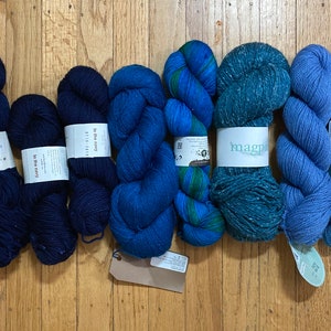 May include: Eight skeins of yarn in various shades of blue, including a multi-colored skein with pink and purple stripes. The yarn is arranged in a row on a wooden surface. The labels on the skeins include the names "Plymouth Blue", "The Blue Mary", "Loft", and "Magpie".