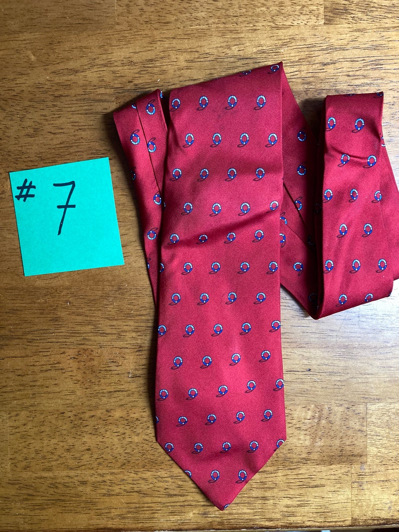 Puede incluir: Una corbata de seda roja con un patr&oacute;n repetido de c&iacute;rculos azules y blancos. La corbata tiene una punta puntiaguda.