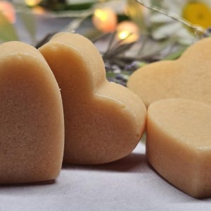 May include: Three solid, light beige, heart-shaped bars of soap arranged together. The soap bars have a smooth, matte texture and are positioned in a cluster, with a single green leaf visible behind the left side of the heart-shaped soap pieces.