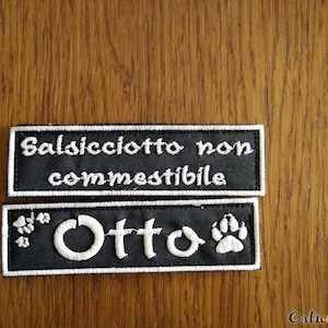 Può includere: Due targhette per cani in tessuto nero con testo ricamato bianco. La targhetta superiore recita "Salsicciotto non commestibile". La targhetta inferiore recita "Otto" con un'impronta di zampa su entrambi i lati del nome.