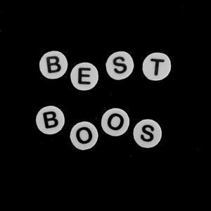 以下が含まれることがあります： BEST BOOSという単語を綴るように配置された、黒い文字入りの白い円形ビーズ。ビーズは無地の黒い背景に対して2列に並べられており、各ビーズにはサンセリフ体のフォントで文字がはっきりと印刷されています。