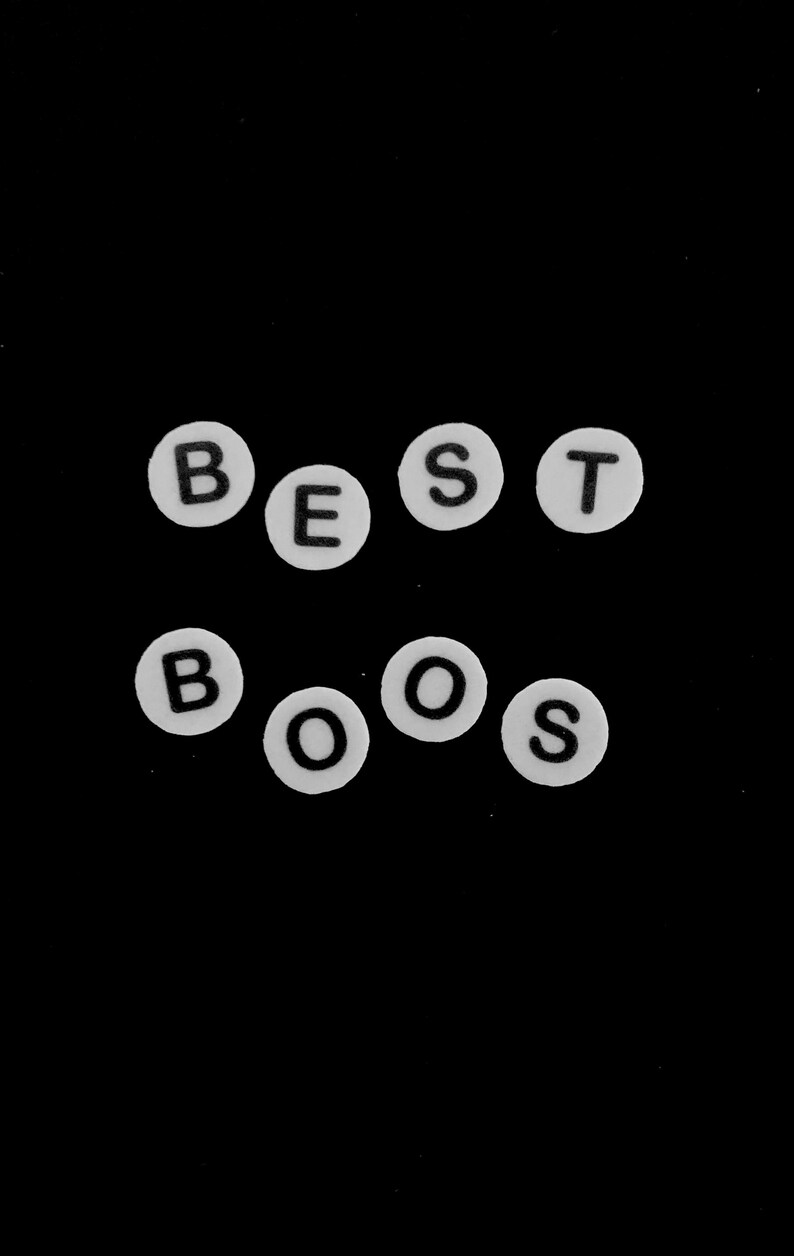 以下が含まれることがあります： BEST BOOSという単語を綴るように配置された、黒い文字入りの白い円形ビーズ。ビーズは無地の黒い背景に対して2列に並べられており、各ビーズにはサンセリフ体のフォントで文字がはっきりと印刷されています。