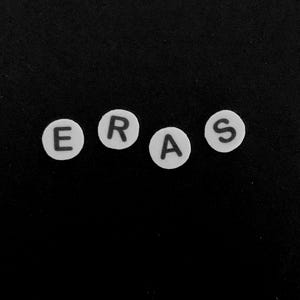以下が含まれることがあります： ERASという単語を綴る、黒い文字が書かれた4つの白い円形のビーズ。各ビーズには、シンプルなサンセリフフォントで大文字が1つずつ印刷されています。ビーズは無地の黒い背景に対して水平に並べられています。