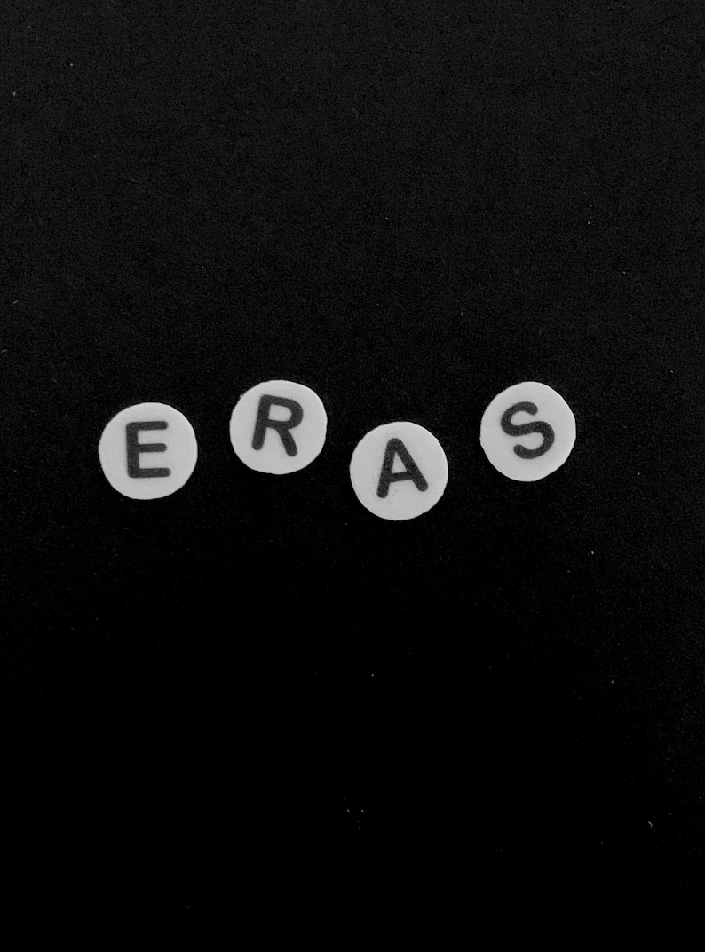 以下が含まれることがあります： ERASという単語を綴る、黒い文字が書かれた4つの白い円形のビーズ。各ビーズには、シンプルなサンセリフフォントで大文字が1つずつ印刷されています。ビーズは無地の黒い背景に対して水平に並べられています。