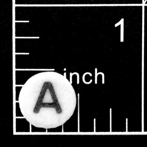 以下が含まれることがあります： 中央に黒い大文字のAがプリントされた、小さく丸い白いボタン。黒い定規の白い目盛りの横に配置されており、この円形のオブジェクトの直径が約1.3センチメートルであることを示しています。