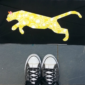 Puede incluir: Un gato de tela amarillo con un patrón floral blanco salta sobre un fondo negro. Dos zapatillas negras y blancas están en primer plano.