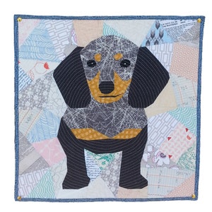 May include: A patchwork quilt featuring a dachshund dog with black and grey fur, a brown nose, and yellow paws. The quilt is made of various fabrics with different patterns and colours, including blue, green, pink, and white.