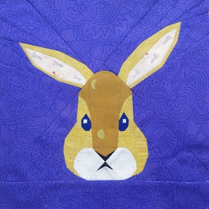May include: A yellow and brown bunny rabbit with white ears and a white nose. The bunny is on a purple background with a floral pattern.
