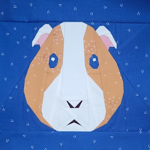 May include: A white and brown patchwork fabric applique of a guinea pig with brown ears and black eyes. The guinea pig is on a blue background with white dots.