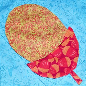 May include: A square quilt block featuring a stylized acorn design. The acorn cap is a yellow and orange floral pattern, while the acorn body is red with orange acorn shapes. The background is light blue with a whimsical pattern.