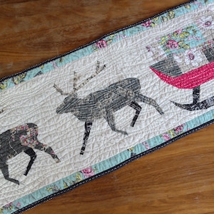 May include: A white quilted table runner with a black border features a festive design of two reindeer and a red sleigh with a floral pattern. The runner is perfect for adding a touch of holiday cheer to your table.