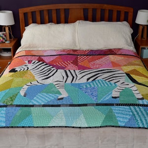 Op de afbeelding: Een kleurrijk quilt met een zebra-ontwerp. Het quilt is gemaakt van verschillende tinten blauw, groen, geel, oranje, rood en roze. De zebra is zwart-wit en bevindt zich in het midden van het quilt. Het quilt ligt op een bed met witte lakens en kussens.