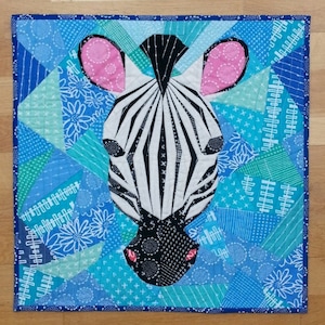 Puede incluir: Un bloque de colcha de cerca de una cabeza de cebra en blanco y negro. La cebra tiene orejas rosas y está sobre un fondo de patchwork azul y verde.