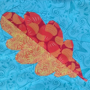 May include: A quilted fabric square featuring a large oak leaf design. The leaf is composed of orange and red fabrics with acorn patterns and yellow floral accents. The background is a light blue fabric with a repeating pattern of squirrels and swirling lines.