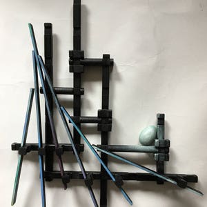 May include: Abstract wall art sculpture made of black and blue painted wooden pieces. The sculpture is composed of a grid of black pieces with blue pieces extending out at various angles. A single blue-green egg-shaped object rests on top of the sculpture.